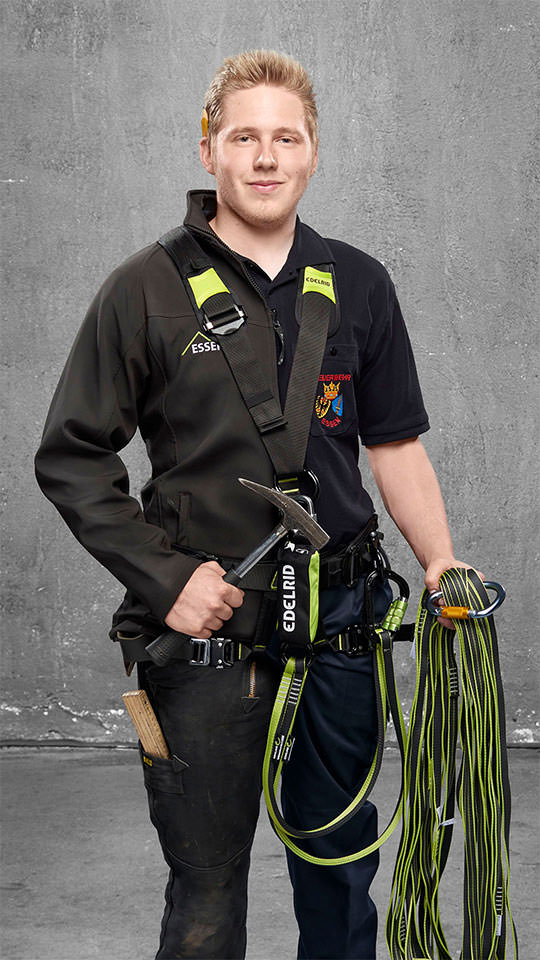 Fotomontage eines Mannes: Halb Dachdecker in schwarzer Arbeitskleidung mit Sicherung für die Höhe, halb Feuerwehrmann in Uniform. Er trägt Bandschlingen und einen Latthammer.