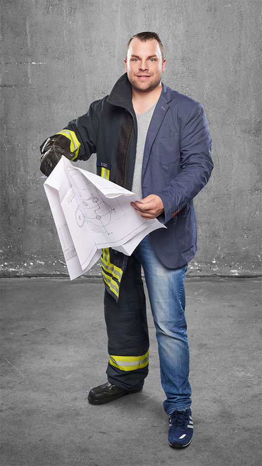 Fotomontage eines Maschinenbauers: Zur einen Hälfte in blauem Sakko und Jeans, zur anderen Feuerwehrmann. Er hält eine technische Zeichnung in den Händen.
